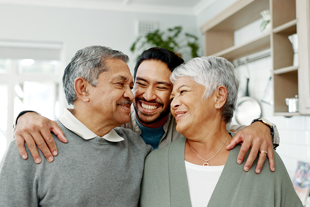 A smiling man hugs an older man and woman in a bright kitchen. All three look happy and close, sharing a joyful moment together. The older couple faces each other, and the man stands between them with arms around their shoulders.