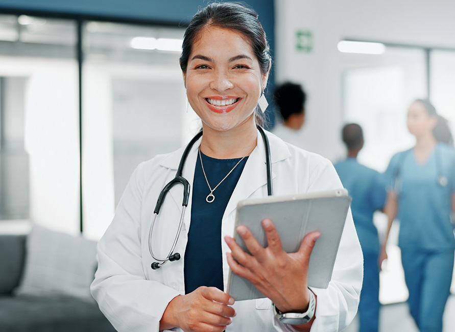 Smiling female doctor holding a tablet in a hospital hallway with medical staff in the background.