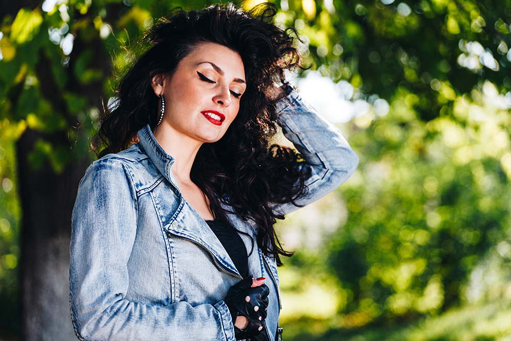 A woman with long dark hair and red lipstick wears a denim jacket and black fingerless gloves, standing outdoors with eyes closed, hand in her hair, and green trees in the background.