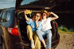 Two young women sit in the open trunk of a car by a lake, smiling and taking a selfie with a yellow phone. They wear casual clothes, sunglasses, and one has a hat. It’s sunny and outdoors.