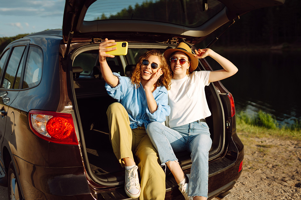 Two young women sit in the open trunk of a car by a lake, smiling and taking a selfie with a yellow phone. They wear casual clothes, sunglasses, and one has a hat. It’s sunny and outdoors.